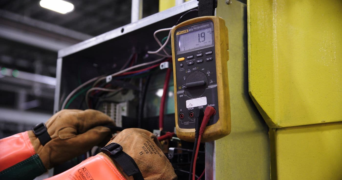 Gloved hands use a Fluke multimeter to take an electrical measurement inside an industrial control cabinet.